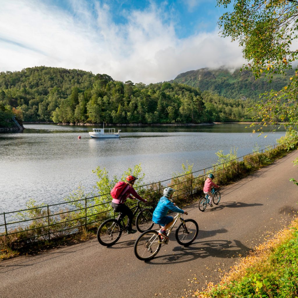 country path with family cycling beside loch katrine and green hills in background